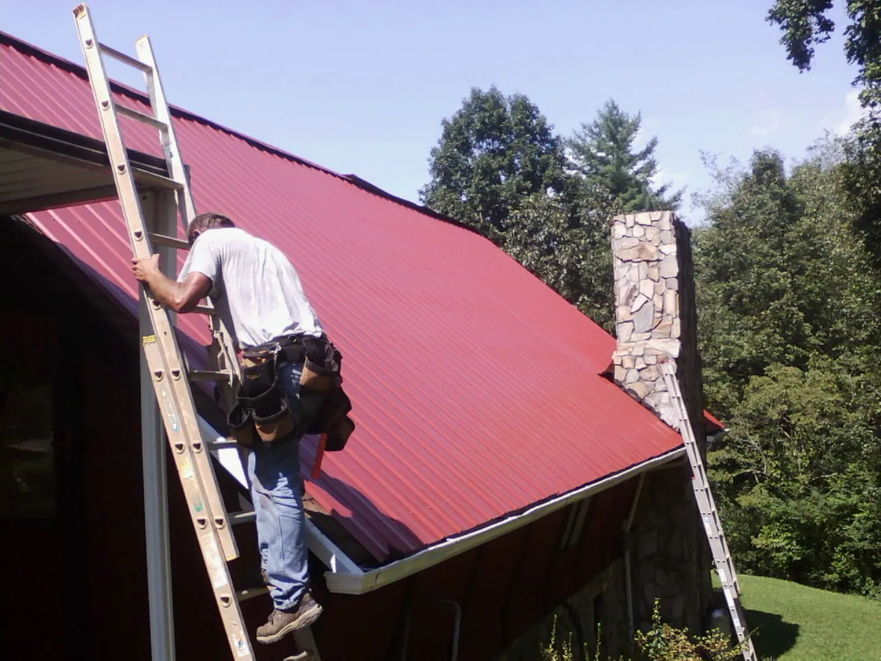 Roofer on a ladder beside a red metal roof and stone chimney — Soffit & Fascia Repair by Roofing Brothers in Dunkirk